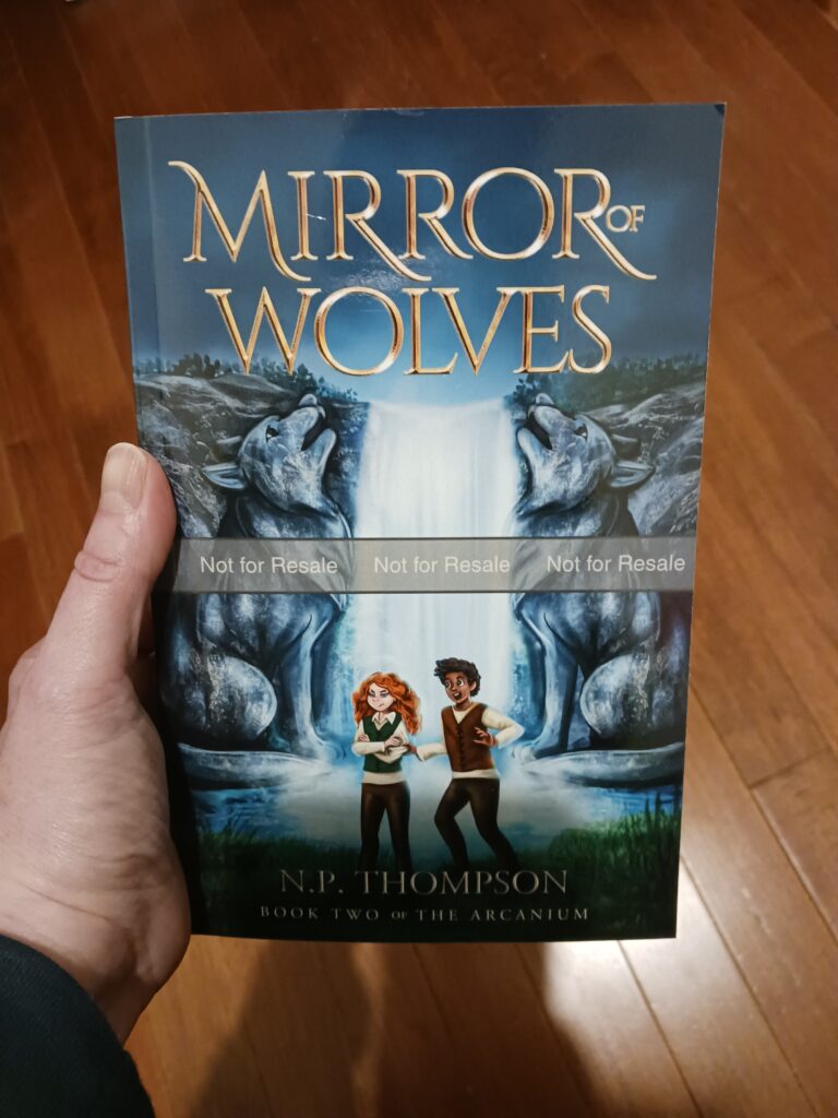 A hand holding a proof copy of Mirror of Wolves, a middle grade fantasy novel. It's a paperback featuring two howling wolves carved into a mountain. A waterfall cascades between them into a pool of water. In the foreground, a boy excitedly yanks on the sleeve of a girl whose arms are crossed. She looks less than impressed. 